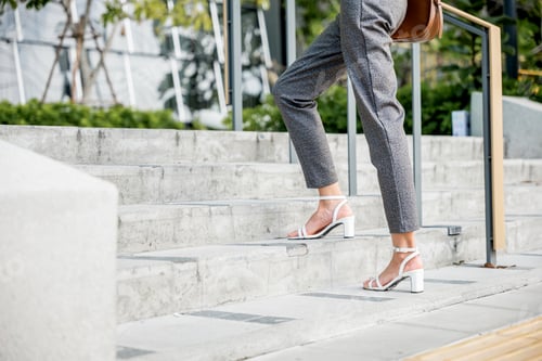 Preview: A businesswoman, portrayed from behind, races up the office stairs, her black shoes stepping towards