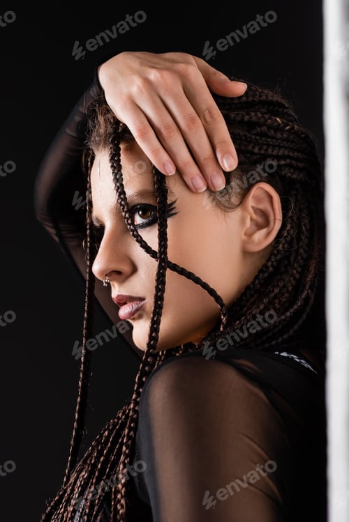 Preview: portrait of stylish woman with makeup and piercing touching braids near white wall on black