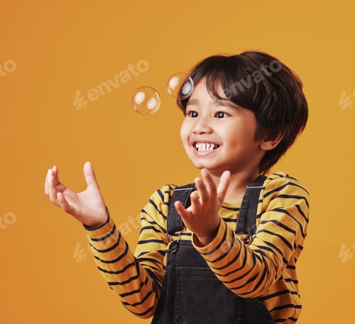 Preview: An adorable little asian boy looking happy while playing with bubbles against an orange copyspace b