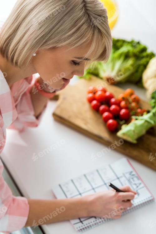 Preview: overhead view of girl writing in notebook with meal lettering near ingredients in kitchen