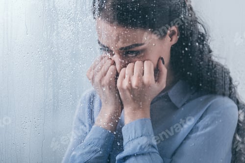 Preview: depressed woman covering face with hands and crying at home through window with raindrops