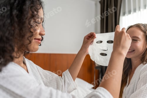 Preview: Two young beautiful woman putting smooth facial mask on face in bedroom.