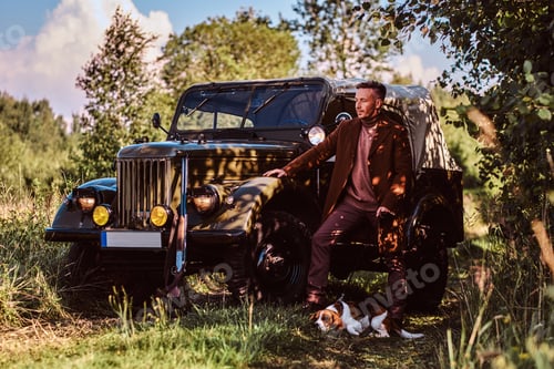 Preview: Old fashioned hunter standing together with his beagle dog near a retro military car in a forest.