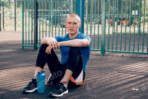 Preview: young man athlete sitting on sports groundr after training, look at camera