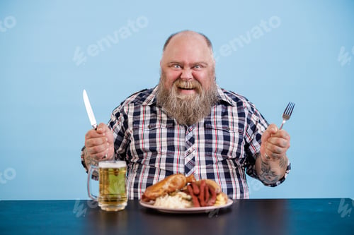 Preview: Joyful person with overweight sits at table with rich food and beer on blue background