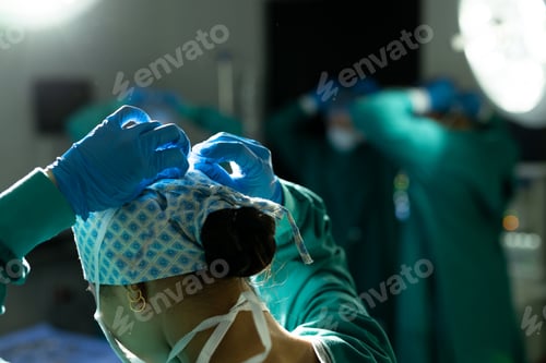 Preview: Rear view of biracial female surgeon in gloves and cap tying mask in theatre, with copy space