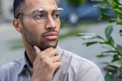 Preview: A close-up photo of a young Hispanic man standing outside the city wearing glasses, rubbing his