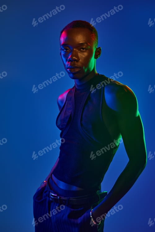 Preview: Young man in stylish attire stands confidently under vibrant blue and green lights