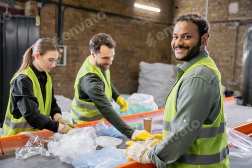 Preview: Joyful indian worker in protective vest and gloves looking at camera while standing near trash