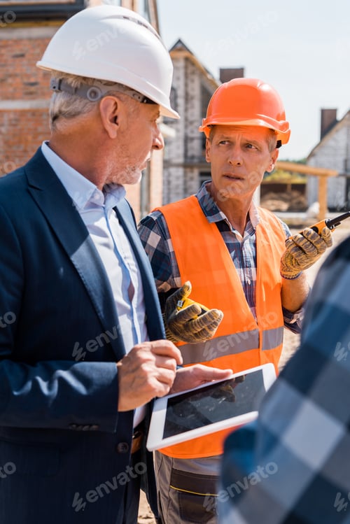 Preview: selective focus of constructor looking at bearded businessman while holding walkie talkie