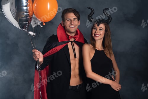 Preview: cheerful man in vampire halloween costume holding balloons near girl with horns on black with smoke