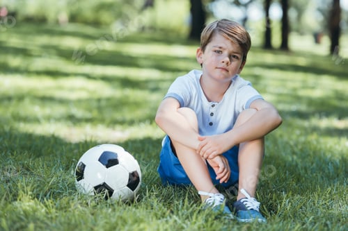 Preview: cute little boy sitting on grass with soccer ball and looking at camera in park