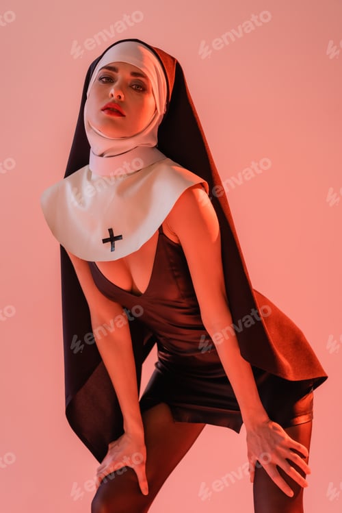 Preview: seductive nun in black leather dress looking at camera isolated on pink
