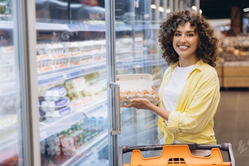 Preview: Woman Holding Eggs in Supermarket Refrigerator Smiling at Camera