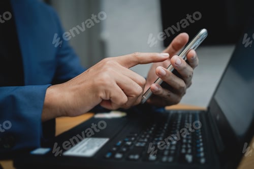 Preview: A man is using a cell phone while sitting at a desk with a laptop.