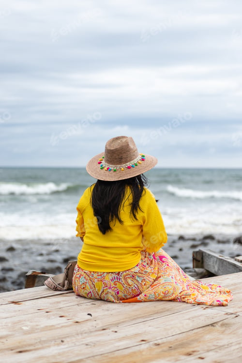 Preview: Contemplative Woman with Straw Hat Sitting on Dock Overlooking the Ocean