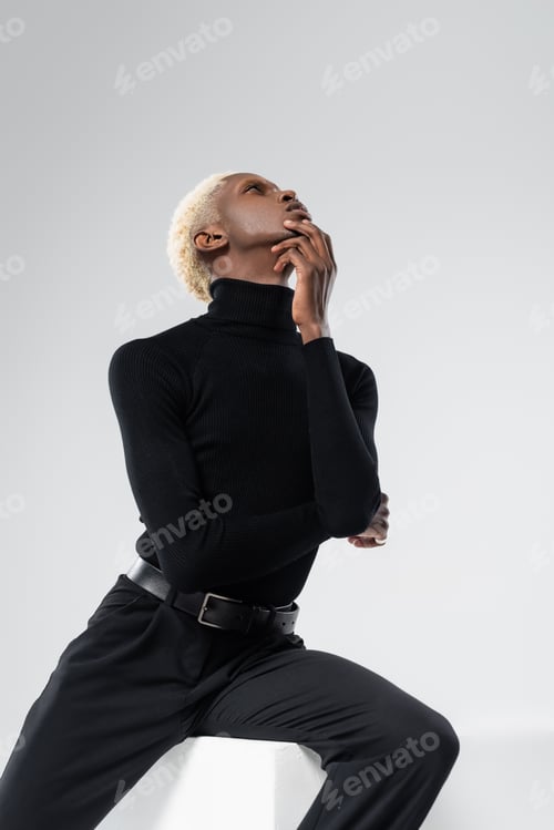 Preview: blonde african american man in turtleneck posing while sitting on white cube isolated on grey