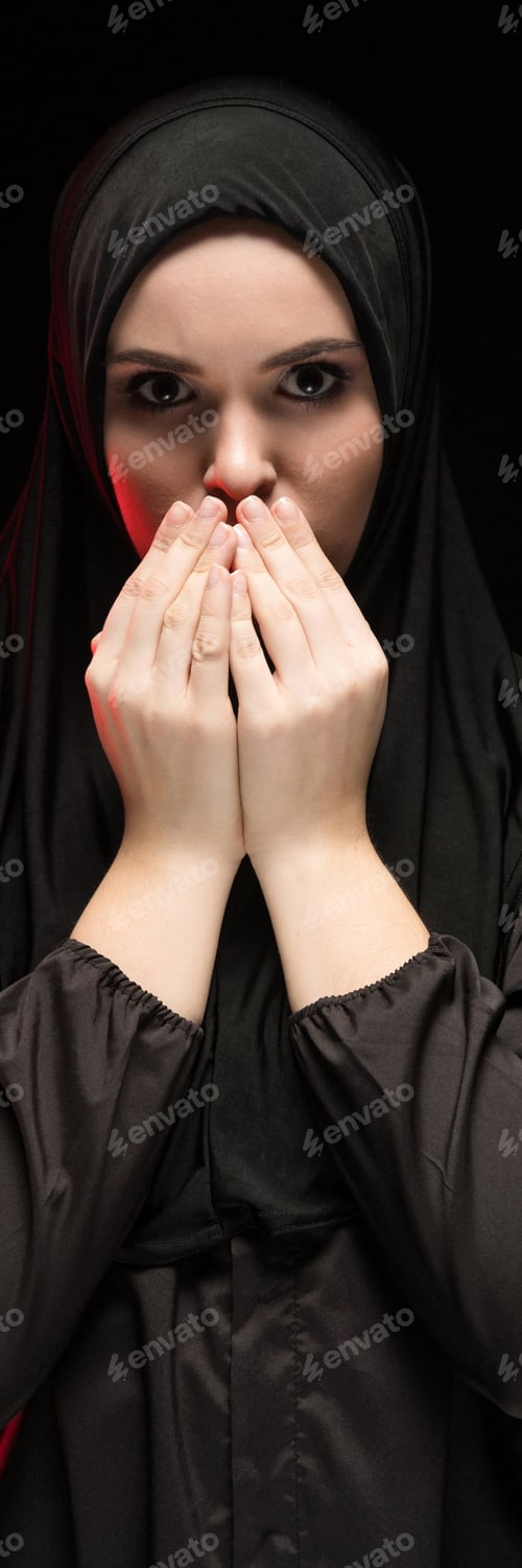 Preview: Portrait of beautiful serious young muslim woman wearing black hijab with hands near her face as