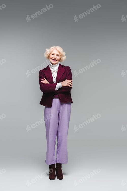 Preview: smiling senior woman with crossed arms in stylish clothes looking at camera on grey