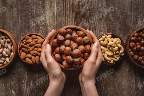 Preview: partial view of woman holding bowl with hazelnuts on wooden tabletop with different nuts around