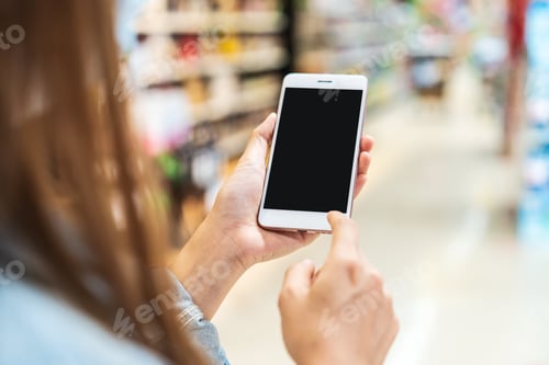 Preview: Young woman holding the empty screen smart phone while shopping in grocery store at the supermarket