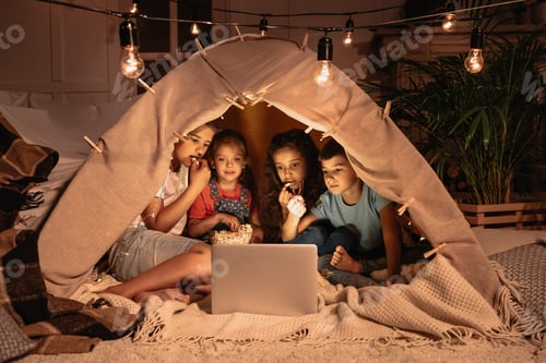 Preview: little multiethnic kids sitting tent and watching film together on laptop