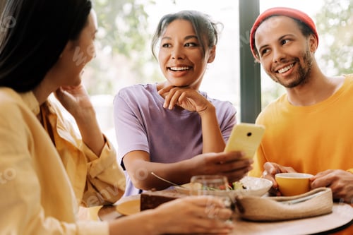 Preview: Happy diverse friends sharing funny content on mobile phone while having lunch together in cafe