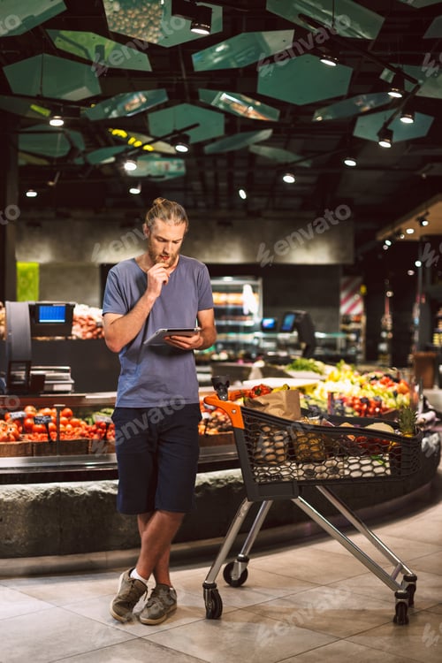 Preview: Young man with trolley full of products thoughtfully using the t