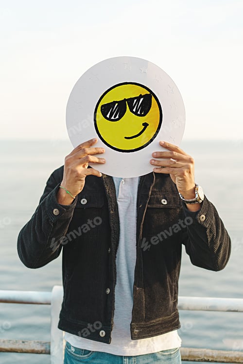 Preview: Young adult man cover face with a smiling emoji face with sunglasses, hand drawn on a white disc