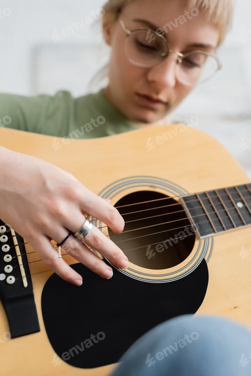 Preview: talanted young woman in glasses with bangs, rings on fingers playing acoustic guitar and sitting