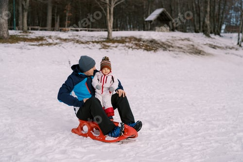 Preview: Smiling dad looking at little daughter sitting on sled on snowy plain