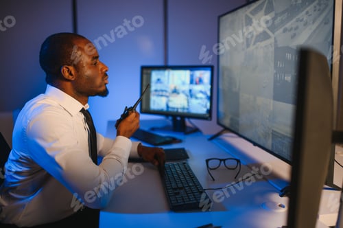 Preview: African American security guard looking at desktop computer screen while speaking on walkie talkie