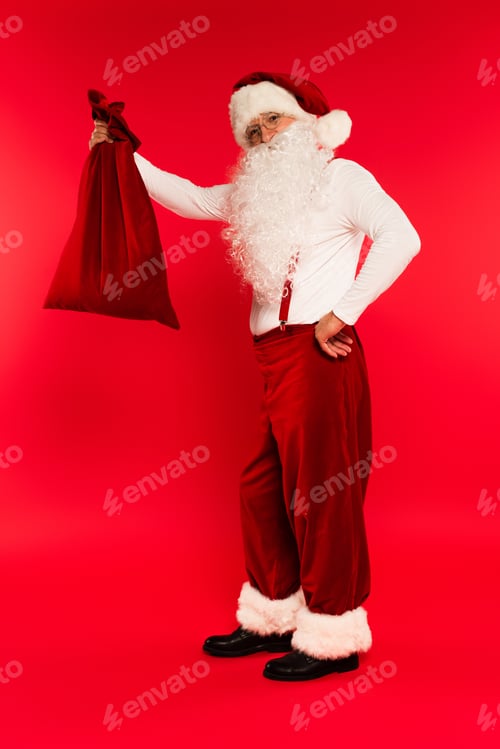 Preview: Full length of bearded santa claus holding sack and looking at camera on red background