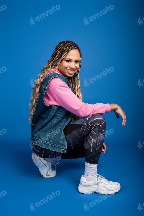 Preview: happy african american woman with dreadlocks posing on blue background, sporty outfit, smile
