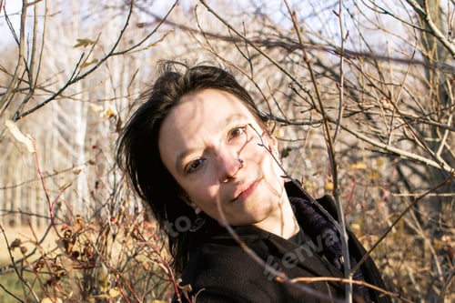 Preview: A young woman with long brown hair wearing a black coat in the middle of some bushes looking down.