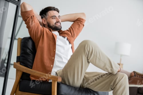 Preview: happy and bearded man sitting on comfortable leather armchair in living room