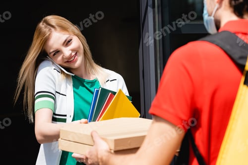 Preview: Selective focus of courier in medical mask giving pizza boxes to smiling girl talking on smartphone