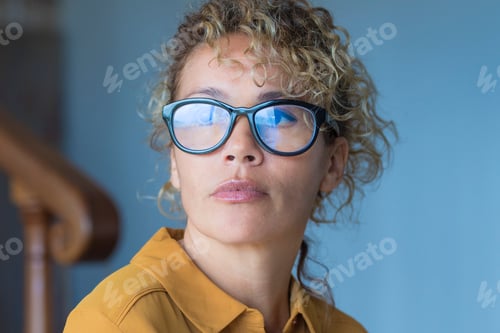 Preview: Portrait of thoughtful young adult woman with eyewear sitting on the sofa at home looking outside