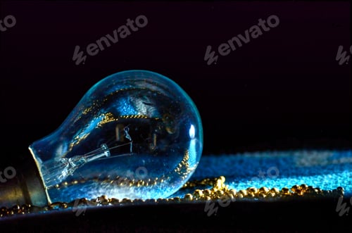 Preview: Closeup shot of aincandescent lamp with blue light and reflecting small golden ball on a black color