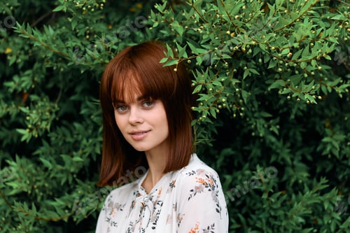 Preview: Vibrant redhead woman posing in front of lush green bush with leaves