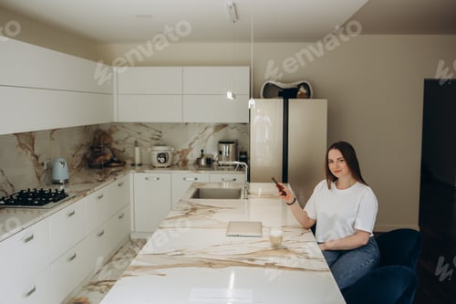 Preview: Young beautiful woman using mobile phone while healthy breakfast in light modern kitchen