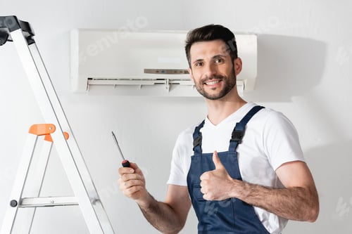Preview: happy repairman showing thumb up and holding screwdriver while standing near stepladder and air
