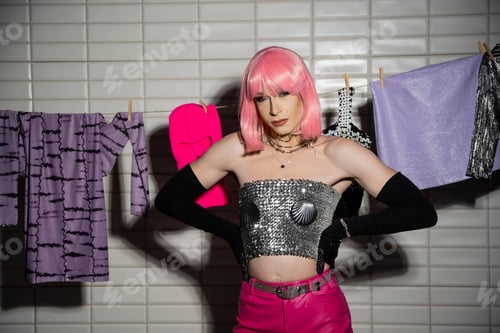 Preview: Stylish drag queen in wig posing near clothes on rope in bathroom