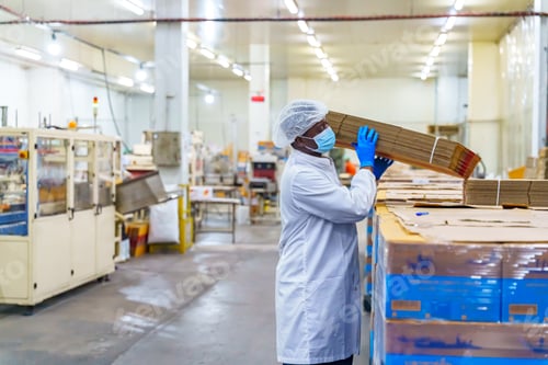 Preview: Factory worker in protective gear carries a stack of cardboard boxes in a warehouse setting