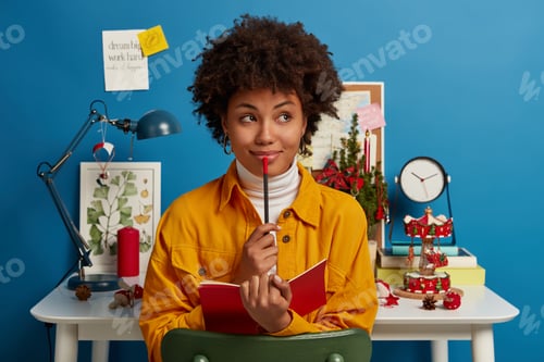 Preview: Pensive dark skinned girl creats interesting composition, sits near workplace with opened notebook a