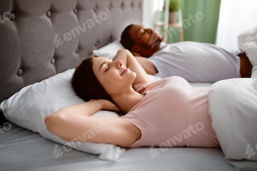 Preview: Happy married interracial couple resting in bed at home, lying with hands behind their heads in
