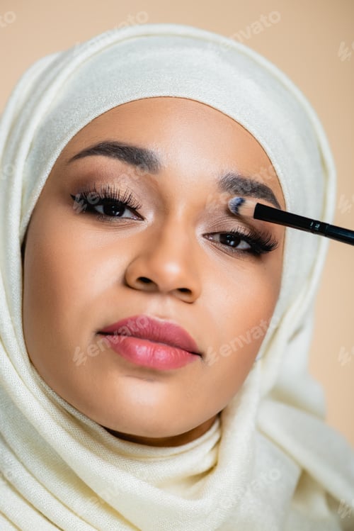 Preview: close up of young muslim woman in hijab applying eye shadow isolated on beige
