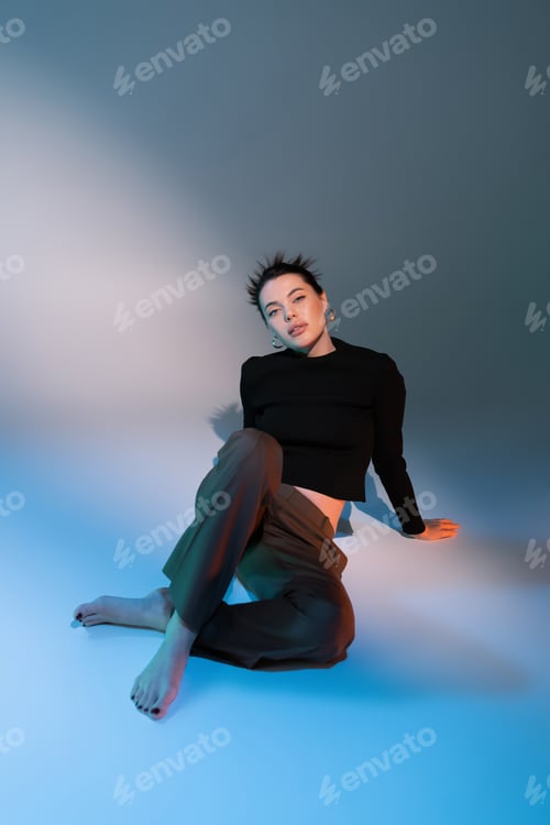 Preview: full length of sensual barefoot woman in brown pants and black sweater sitting on gray and blue
