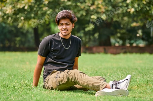 Preview: Smiling young indian man candid portrait in black t shirt, silver neck chain sitting on green lawn