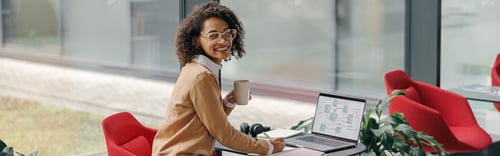 Preview: Business woman in eyeglasses drinking coffee during working on laptop and making notes in cozy cafe